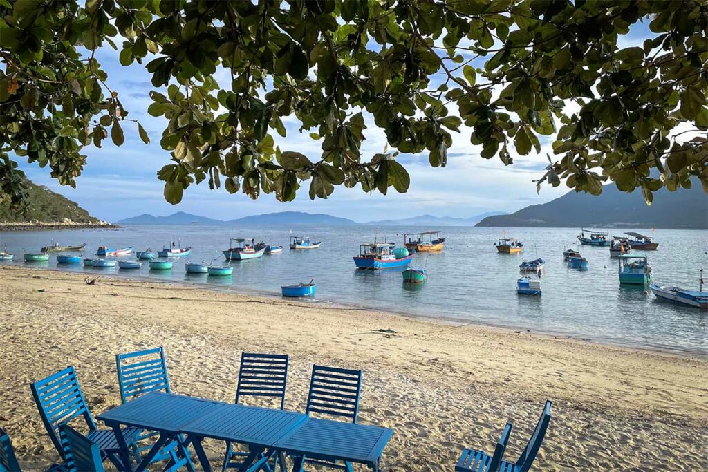 Strand van het vissersdorp Ninh Van met blauwe stoelen en tafels onder een boom, tientallen ronde en houten visserboten in het ondiepe water en bergen aan de horizon