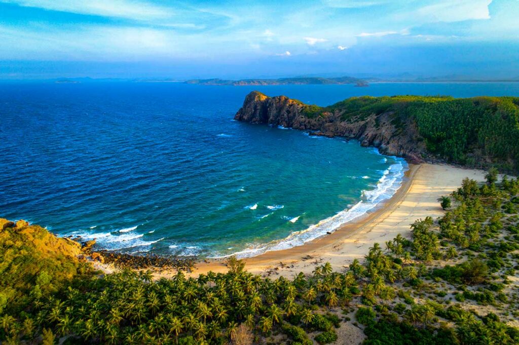 Verlaten halfcirkelvormig strand in Phu Yen met goudgeel zand, blauwe golven, palmbomen en een rotsachtige kaap op de achtergrond — een van de weinig bekende stranden langs de kust van Phu Yen