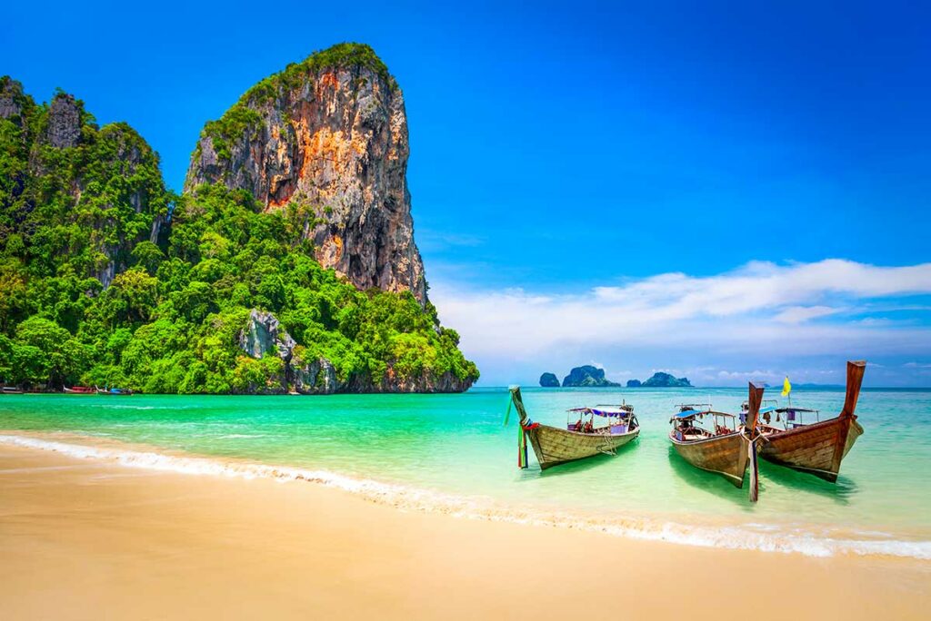 Drie traditionele longtailboten op het heldere turquoise water van Railay Beach in Thailand, met een grote begroeide kalksteenrots op de achtergrond