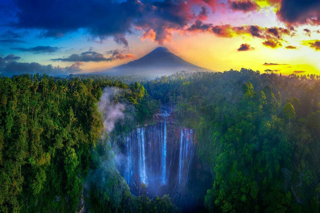 Luchtfoto van de Tumpak Sewu waterval op Java in Indonesie die in een beboste kloof stort, met de conische top van de Semeru vulkaan zichtbaar in de achtergrond bij zonsondergang