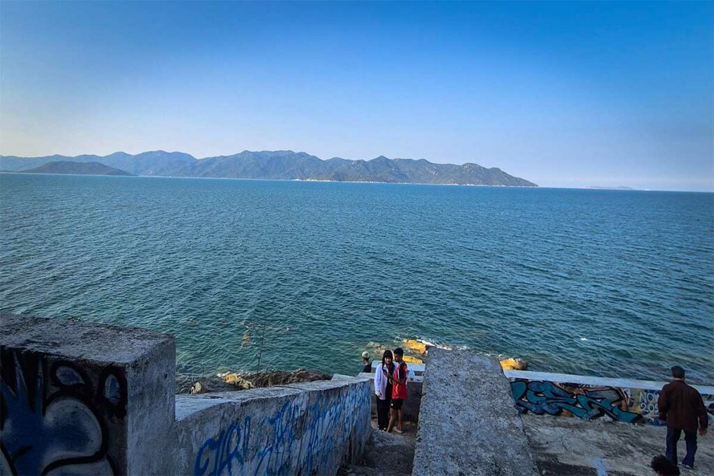 Uitzichtpunt aan de kust van Ninh Van Bay met bezoekers op betonnen trappen met graffiti, uitzicht over de donkerblauwe zee en de beboste bergkam van Ninh Van op de achtergrond