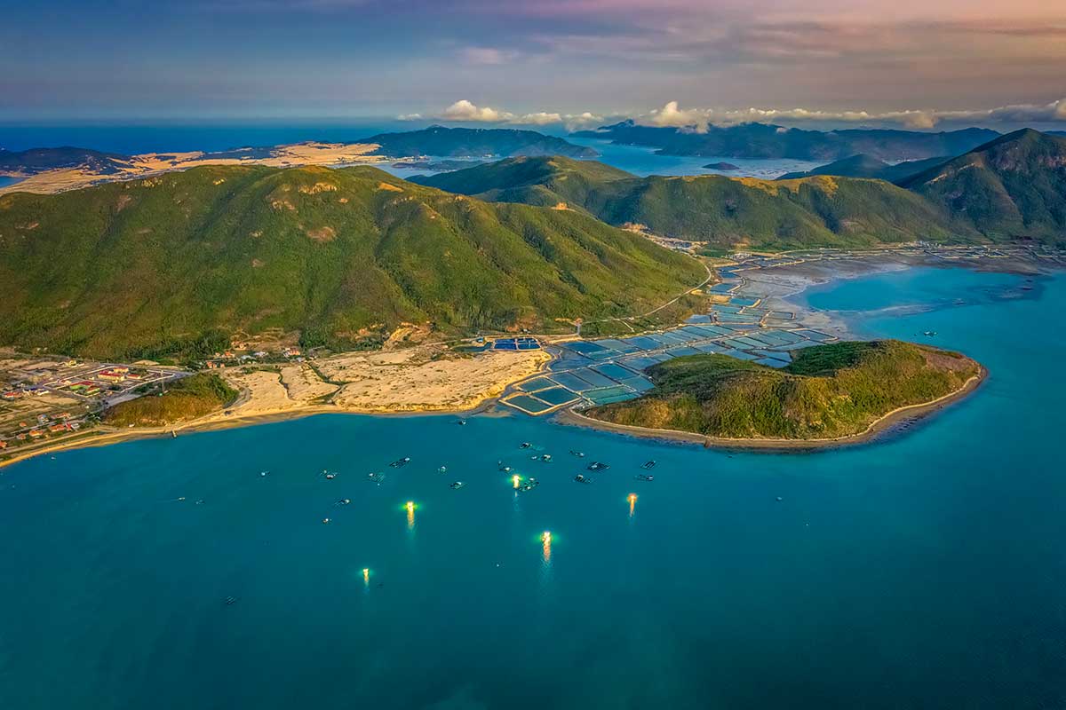 Luchtfoto van de Van Phong Bay bij avondlicht met groene berghellingen, viskwekerijen langs de kust en vissersbootjes met gele lichten op het turquoise water