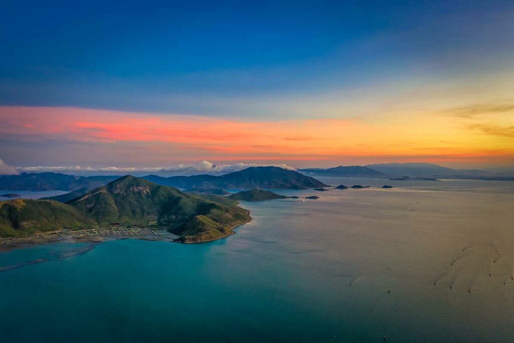 Luchtfoto van de Van Phong Bay bij zonsondergang met een kleurrijke lucht in roze, oranje en blauwe tinten, groene bergeilanden en rustig water — een van de mooiste baaien ten noorden van Nha Trang