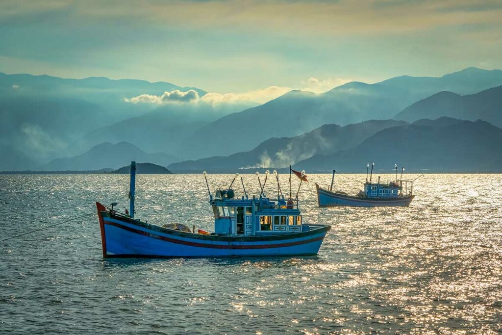 Twee traditionele Vietnamese vissersboten op het glinsterende water van de Van Phong Bay met bergkettingen in de mist op de achtergrond — een authentiek beeld van het vissersleven in de baai