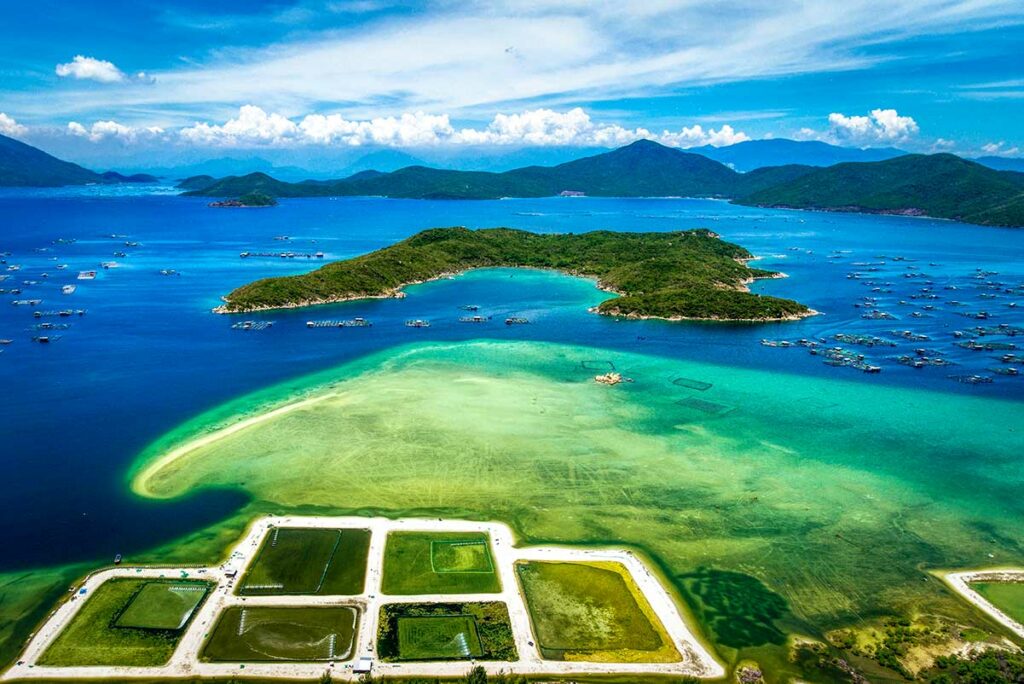 Luchtfoto van de Van Phong Bay met Hon Lon eiland centraal in beeld, omgeven door turquoise en diepblauw water, viskwekerijen en meerdere kleinere eilanden verspreid over de baai