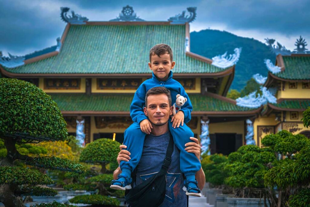 Vader met kind op de schouders voor een Vietnamese tempel tijdens een gezinsvakantie in Vietnam, met groene bonsaibomen en bergen op de achtergrond