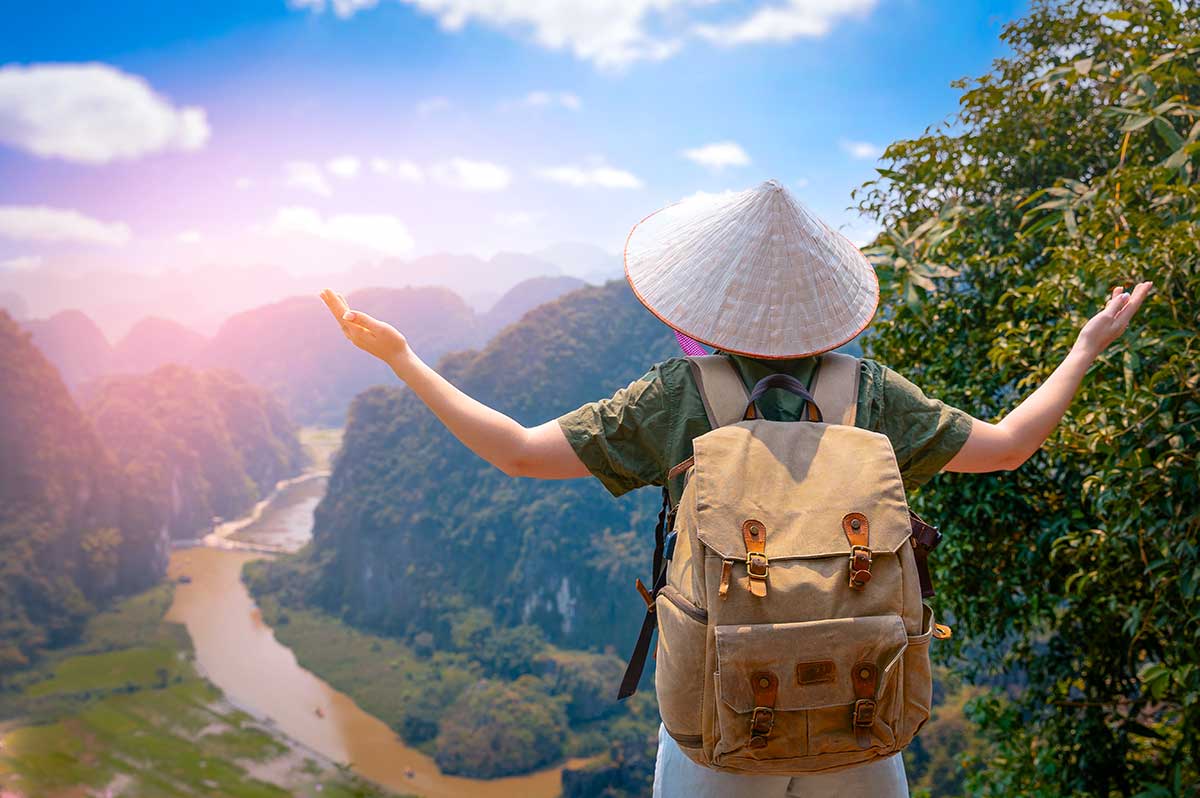 Reiziger met Vietnamese conische hoed en rugzak spreidt de armen uit boven een rivier en kalkstenen bergen in Ninh Binh, een van de hoogtepunten van een rondreis door Vietnam
