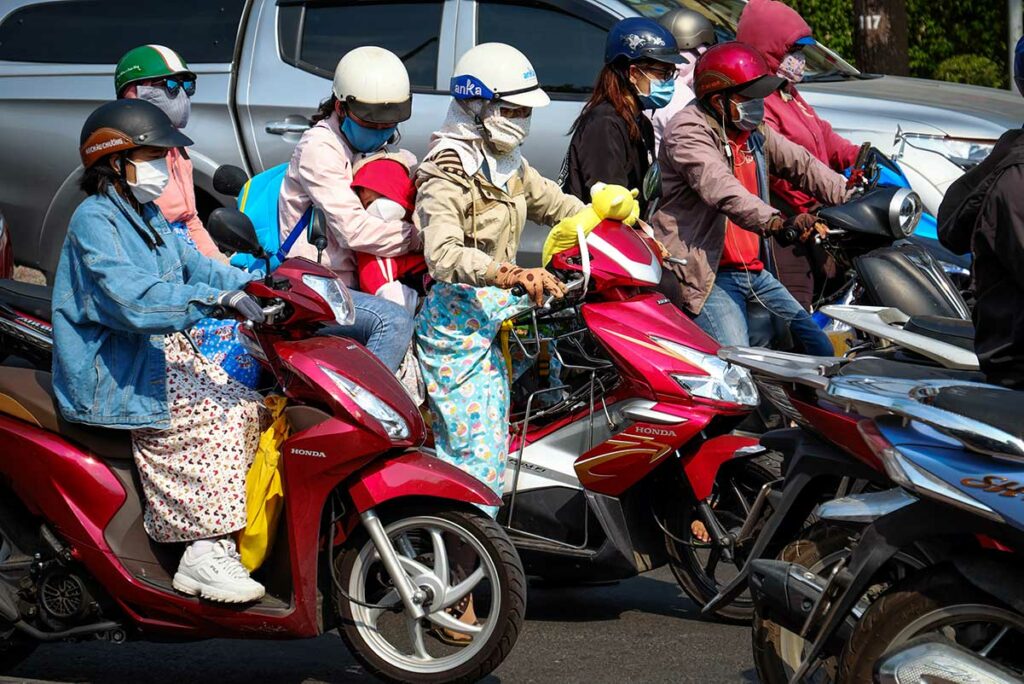 Vietnamezen op scooters bij een druk kruispunt met volledige zonbescherming, waaronder gezichtsmaskers, handschoenen en lange mouwen