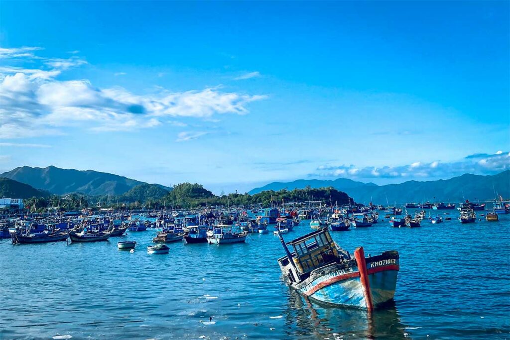 Drukke vissershaven van Ninh Van Bay met honderden blauwe visserboten op het water, een versleten boot op de voorgrond en beboste heuvels op de achtergrond