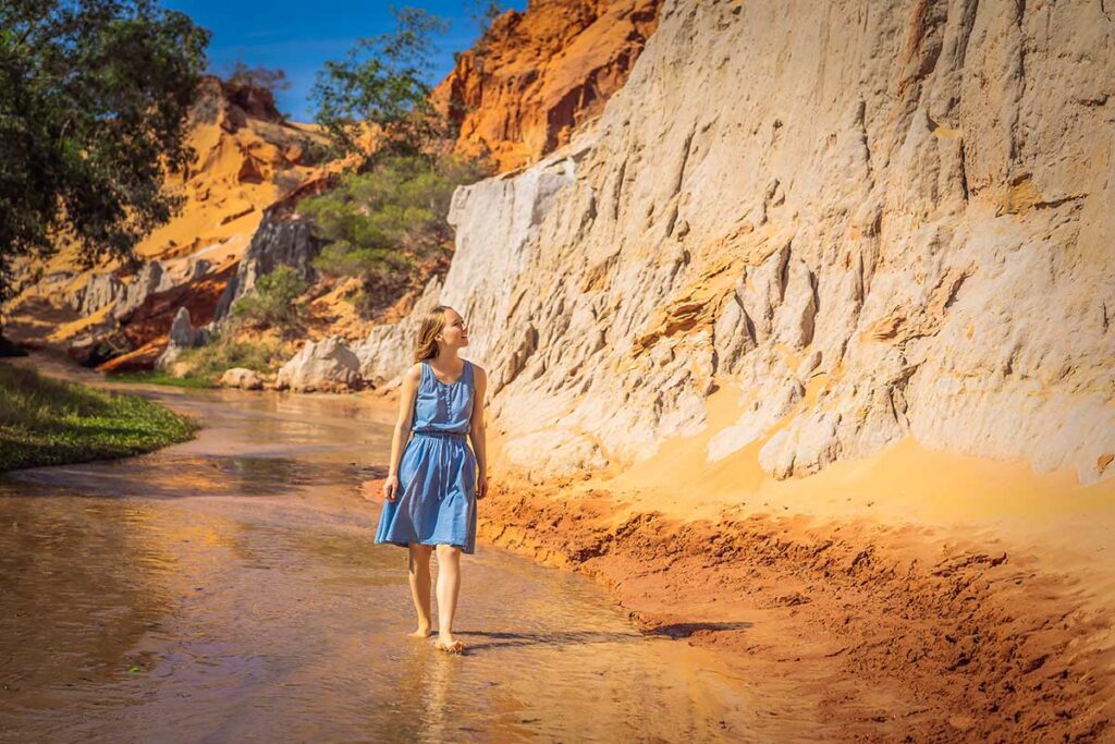 Vrouw in blauwe jurk loopt barrevoets door het ondiepe water van de Fairy Stream in Mui Ne langs de hoge witte zandsteenwand