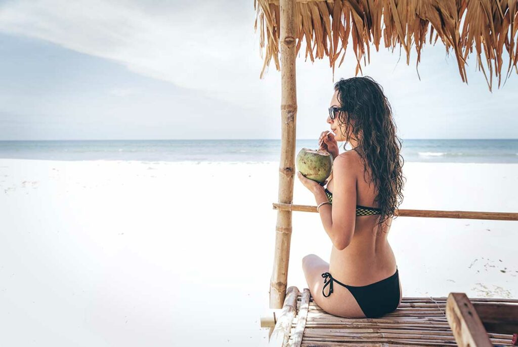 Vrouw in zwart bikini zit onder een bamboese strandhut op een wit zandstrand in Vietnam en drinkt uit een verse kokosnoot met een zonnebril op