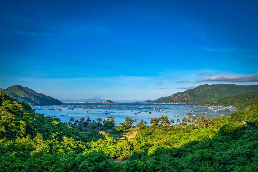 Panoramisch uitzicht over Vung Ro Bay in Phu Yen met helder blauw water, viskwekerijen en visserboten verspreid over de baai, omgeven door dicht beboste heuvels — een afgelegen baai vlak bij de grens met Khanh Hoa