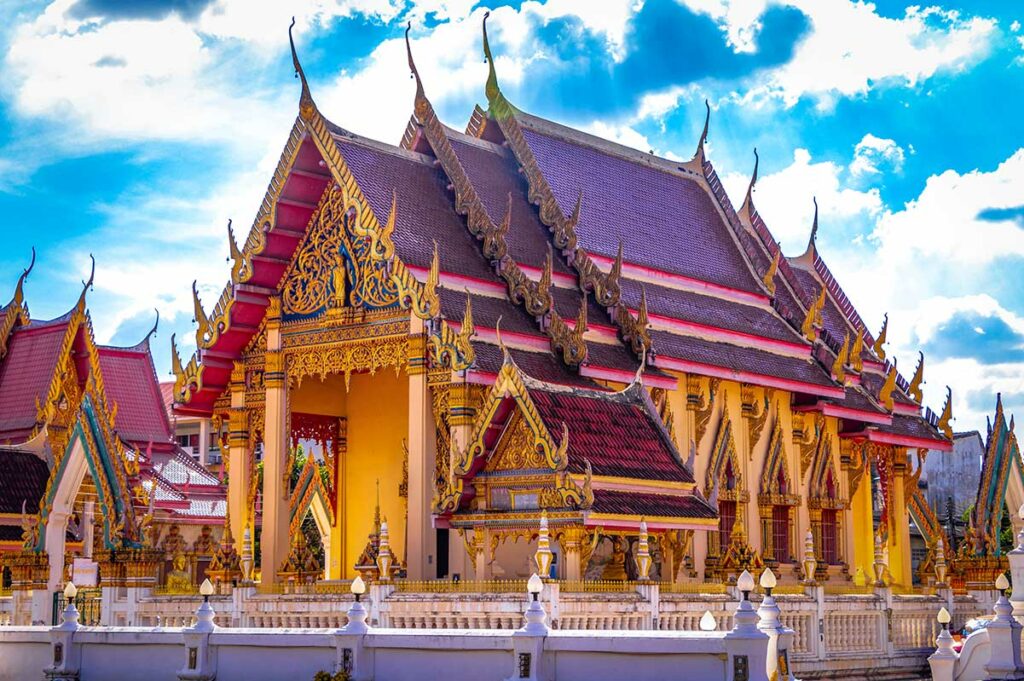 Thaise boeddhistische tempel met gouden ornamenten, rood dak en witte muren onder een blauwe lucht met wolken