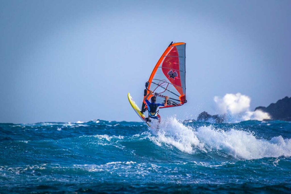 Windsurfer rijdt op een golf in volle zee met een oranje-rode zeil en rotsen op de achtergrond — naast kitesurfen is windsurfen een van de watersportopties in Mui Ne