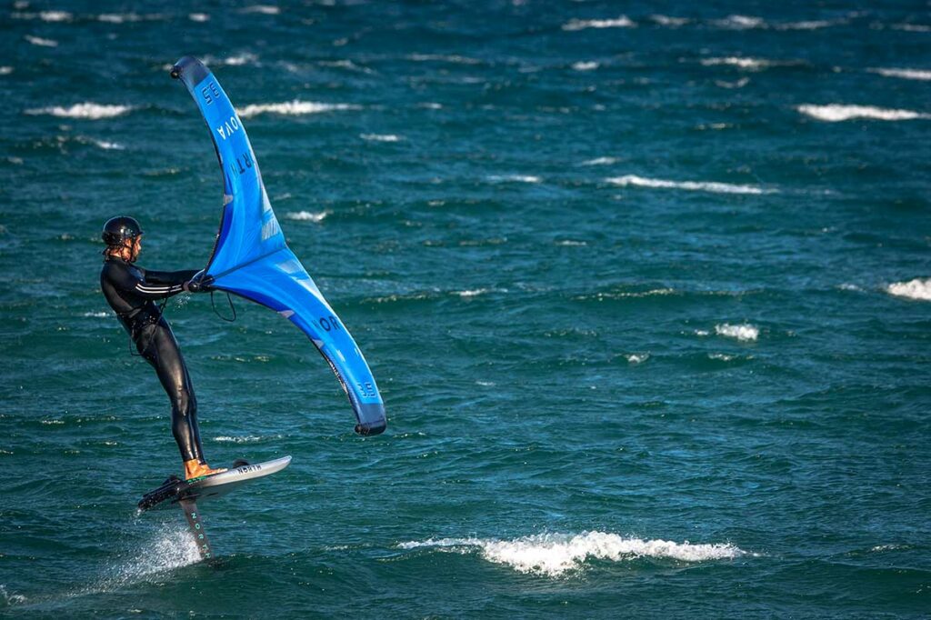 Wing foiler zweeft boven het wateroppervlak op een foilboard met een blauwe vleugel in zijn handen — wing foilen is een nieuwere watersport die ook in Mui Ne beoefend wordt
