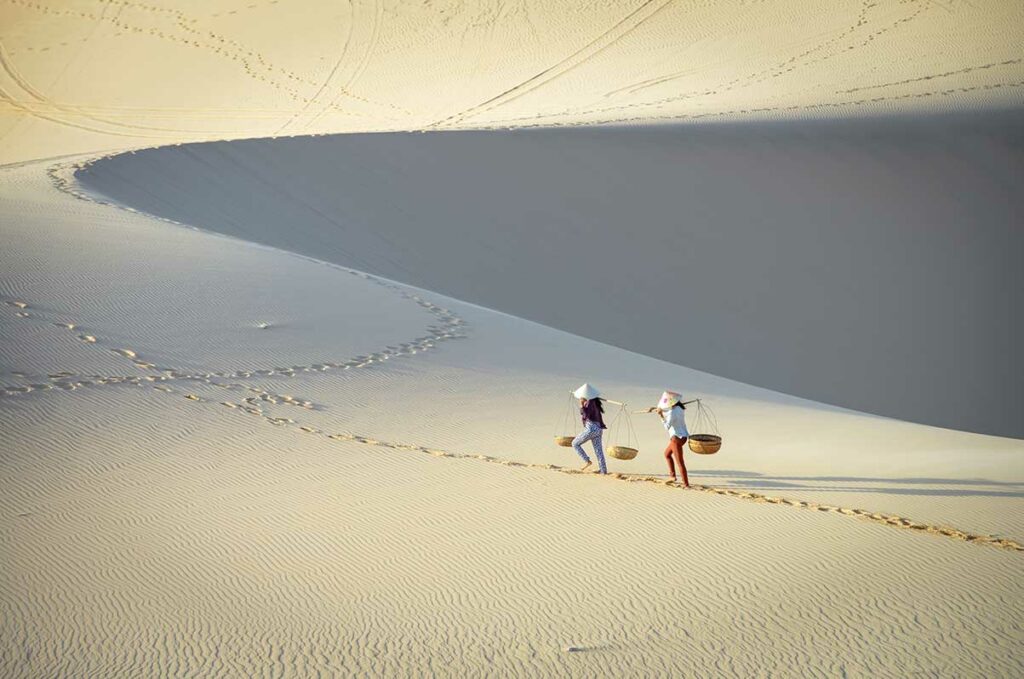 Twee Vietnamese vrouwen met conische hoeden en draagmanden lopen over de uitgestrekte witte zandduinen van Mui Ne — een van de opvallendste natuurlandschappen van Vietnam