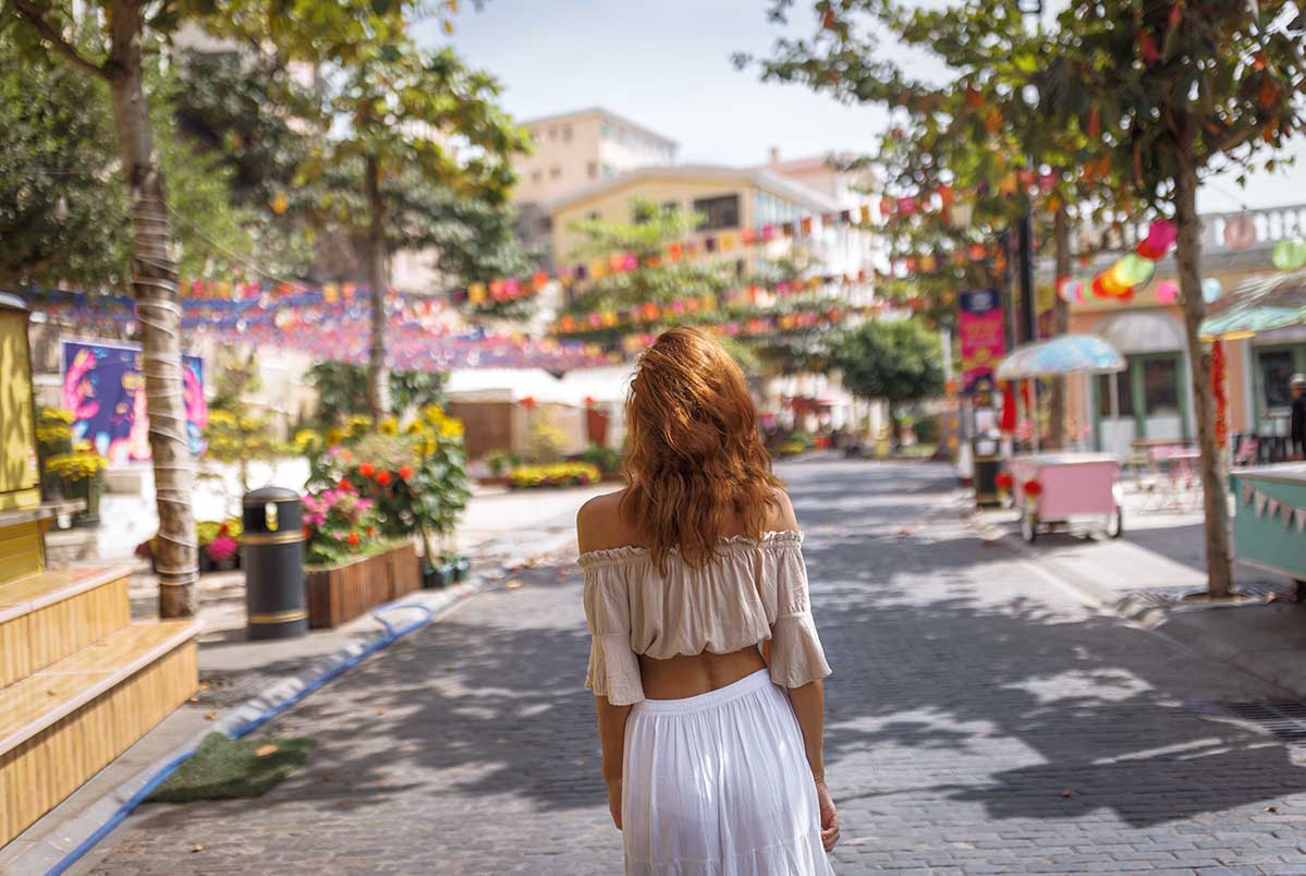 Vrouw in wit off-shoulder outfit loopt door een zonnige straat in Vietnam met kleurrijke decoraties en tropische bomen