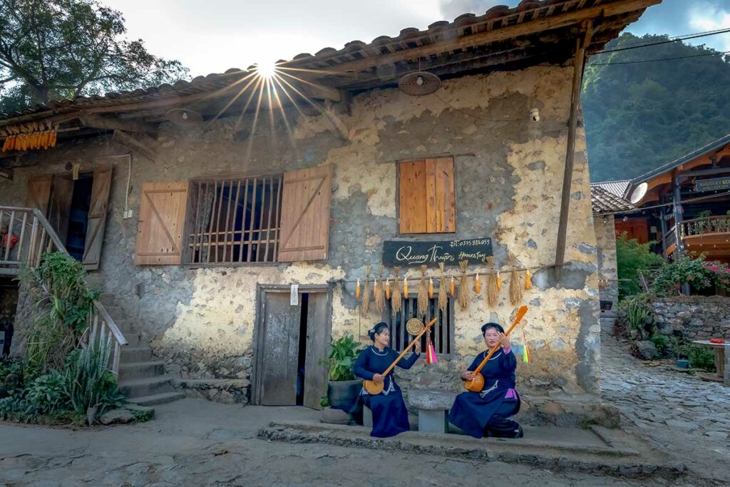 Twee vrouwen in traditionele Tay-kleding spelen snaarinstrumenten voor een stenen huis in Khuoi Ky, Cao Bang