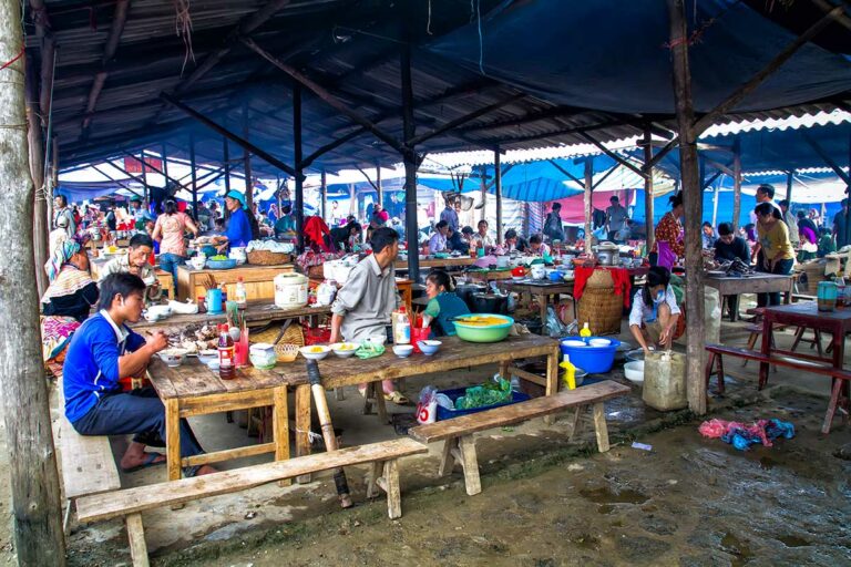 Bezoekers eten aan houten tafels in het overdekte eetgedeelte van de markt van Bac Ha.