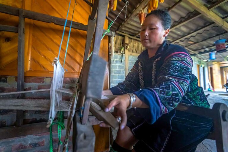 Vrouw weeft aan een houten weefgetouw in een huis in Lao Chai, met maiskolven aan het plafond.