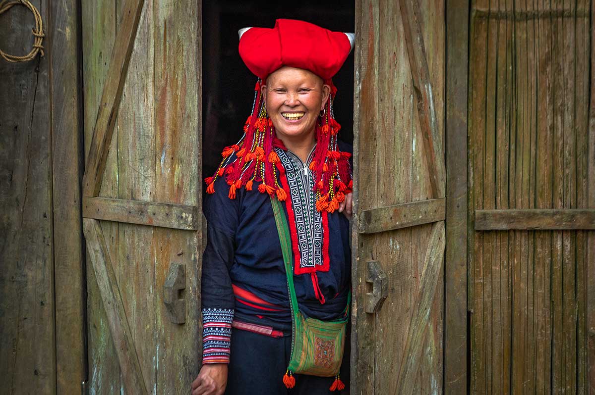 Rode Dao vrouw met rode hoofdtooi lacht in de deuropening van een houten huis bij Sapa.