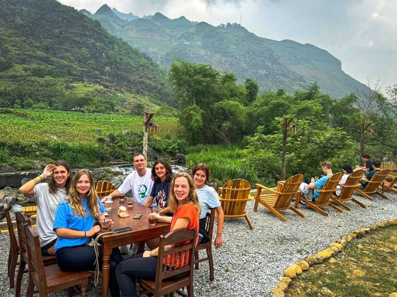 Travelers enjoying drinks at a riverside café in Du Già village surrounded by green mountains.