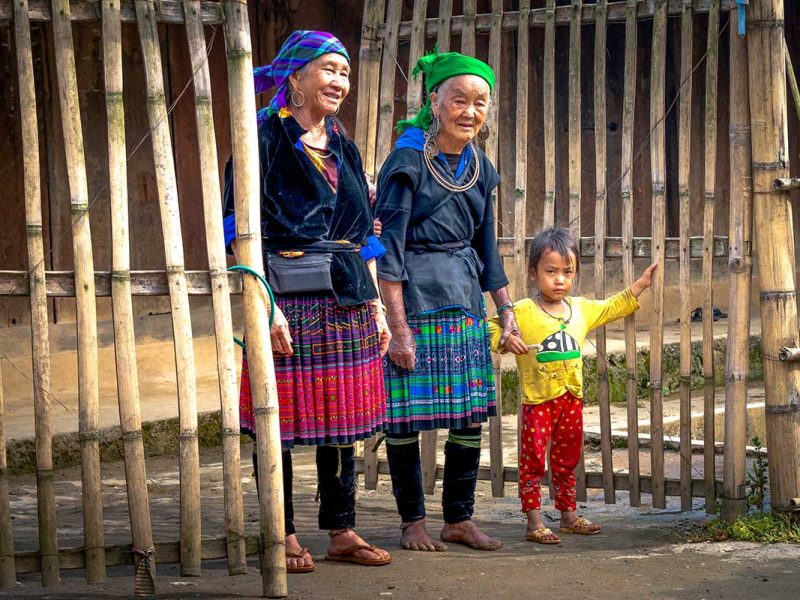 Twee oudere vrouwen in traditionele kleding met een kind bij een bamboehek in Mu Cang Chai