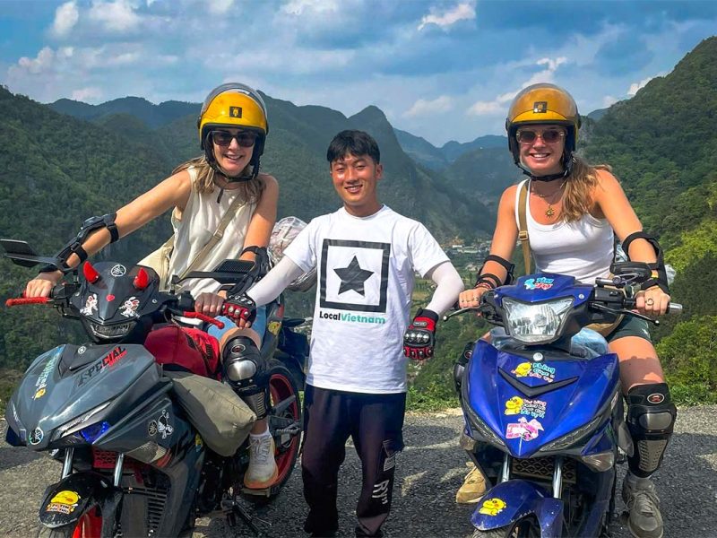 Reizigers poseren met hun Local Vietnam-gids tijdens een Ha Giang Loop Easy Rider tour, met mistige bergpieken en een steile vallei op de achtergrond.