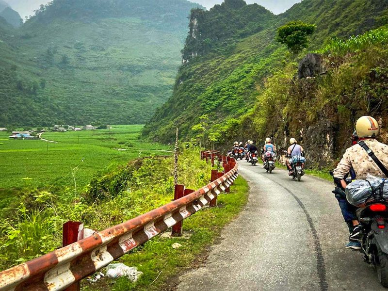 Een groep reizigers rijdt met hun Easy Rider-gidsen door een groene vallei tijdens de Ha Giang Loop groepstour, over een smalle weg langs rotswanden en rijstvelden.