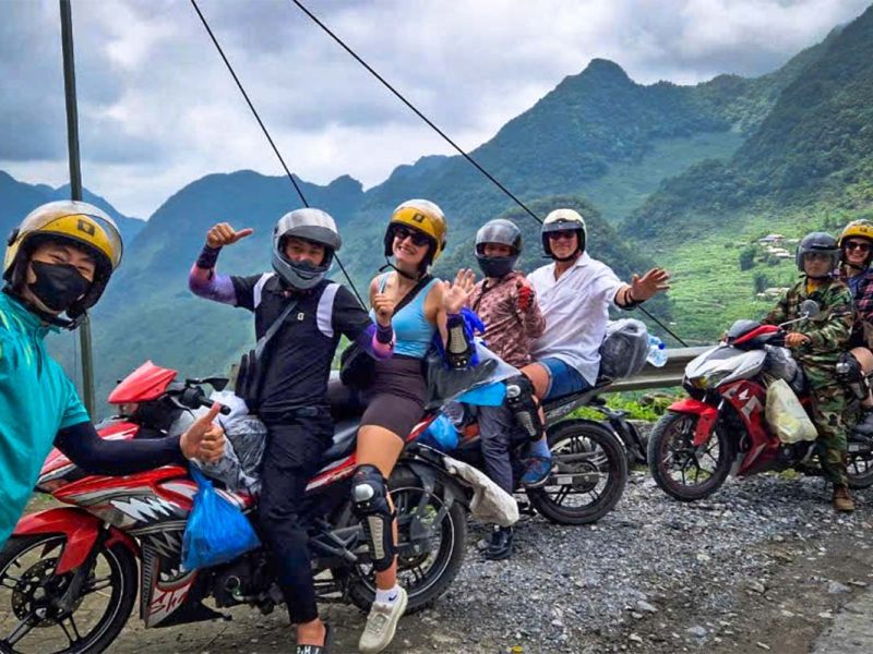 Een groep reizigers op de Ha Giang Loop poseert met hun Easy Rider-chauffeurs langs een bergweg, met ruige bergen en vrolijke groepssfeer op de achtergrond.