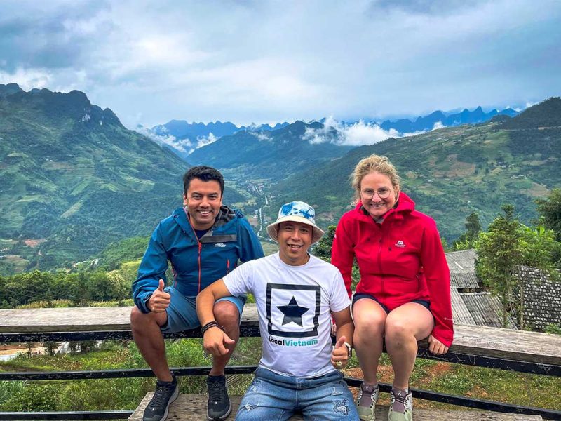 Een Local Vietnam-gids en twee reizigers zitten samen op een houten bankje met panoramisch zicht op een groene vallei en bergtoppen in Ha Giang.