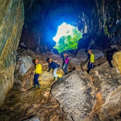 Trekkinggroep met helmen loopt door Elephant Cave in Hang Ma Valley met grote rotsblokken, grotopening en natuurlijk licht in de achtergrond