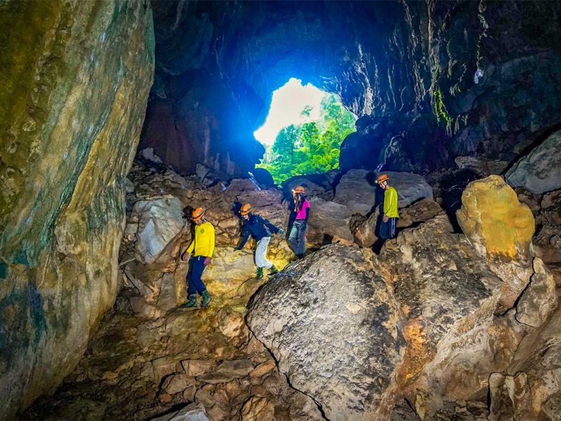 Trekkinggroep met helmen loopt door Elephant Cave in Hang Ma Valley met grote rotsblokken, grotopening en natuurlijk licht in de achtergrond