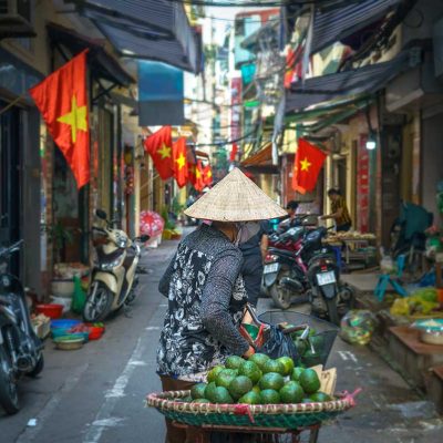 Straatverkoper met conische hoed in een smalle steeg in Hanoi, met meerdere Vietnamese vlaggen die boven de straat hangen.