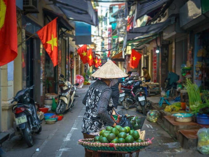 Straatverkoper met conische hoed in een smalle steeg in Hanoi, met meerdere Vietnamese vlaggen die boven de straat hangen.