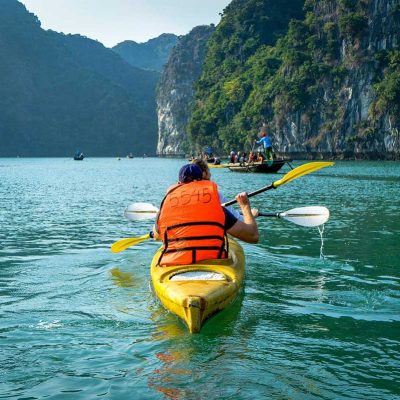 Reiziger in een gele kajak peddelt door Lan Ha Bay, met traditionele bootjes en hoge kalkstenen rotsen langs het kalme water bij Cat Ba Island