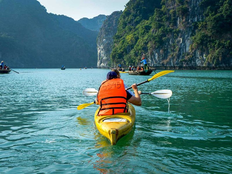 Reiziger in een gele kajak peddelt door Lan Ha Bay, met traditionele bootjes en hoge kalkstenen rotsen langs het kalme water bij Cat Ba Island