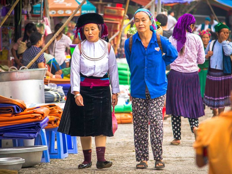 Twee vrouwen in traditionele kleding lopen over de markt in Meo Vac, omringd door kraampjes en textiel.