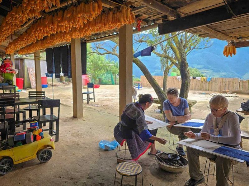 Reizigers tekenen met was op stof onder een afdak met drogende maiskolven in Mu Cang Chai