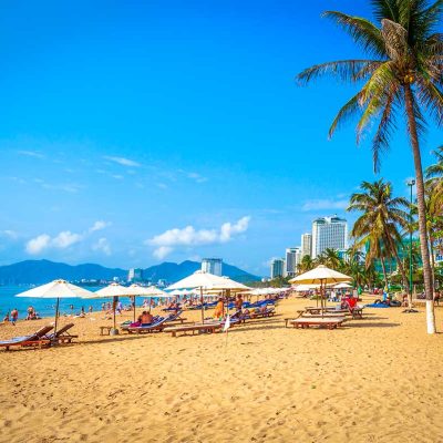 Strand in Nha Trang met ligbedden en parasols langs de zee, palmbomen op de achtergrond en toeristen die ontspannen aan het strand