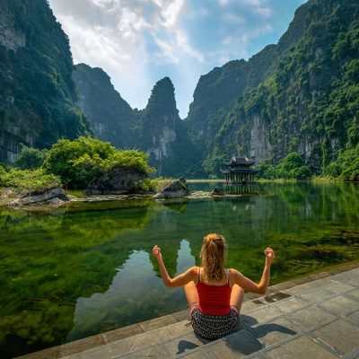 Reiziger zit aan de rand van een pad bij Trang An in Ninh Binh, omringd door groene kliffen en helder water, een rustig moment voor reisfotografie in Vietnam.